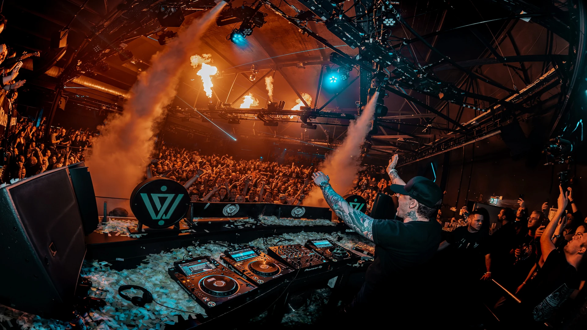 Niels Van Gogh standing behind a DJ Booth with his hands in the air. Fire from pyrotechnics and fog from machines are visible above the crowd.
