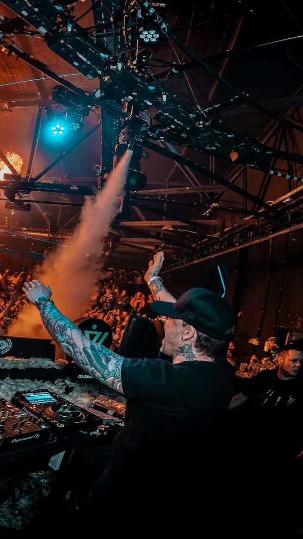 Niels Van Gogh standing behind a DJ Booth with his hands in the air. Fire from pyrotechnics and fog from machines are visible above the crowd.