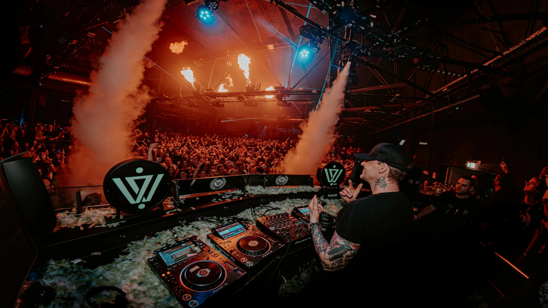 Niels Van Gogh is standing behind a DJ Booth while looking at the crowd and dancing.