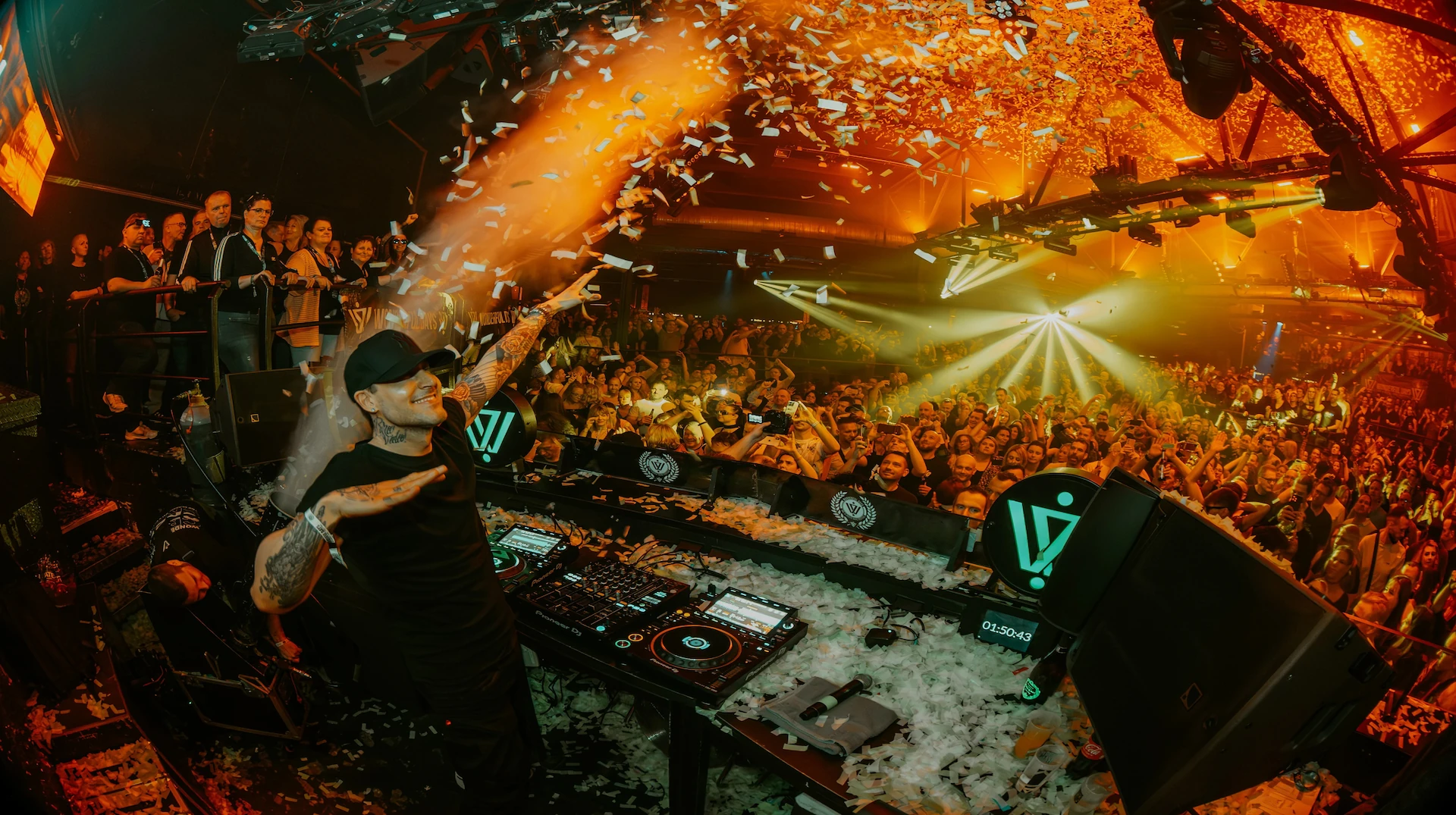 Niels Van Gogh is dancing behind a DJ booth in front of a crowd. There is fire from pyrotechnics above the crowd and its raining confetti.