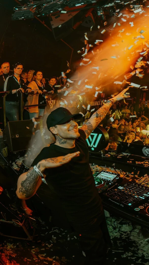 Niels Van Gogh is dancing behind a DJ booth in front of a crowd. There is fire from pyrotechnics above the crowd and its raining confetti.